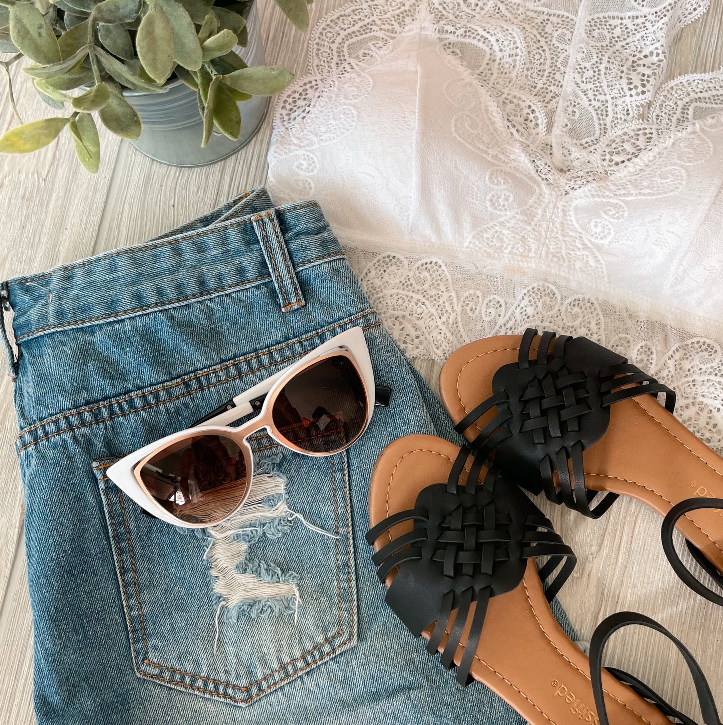 Flat lay of a summer outfit with distressed denim shorts, a white lace bralette, black woven sandals, and Katrina Sunnies • Adult for eye protection, styled beside a green potted plant on a light wood surface.