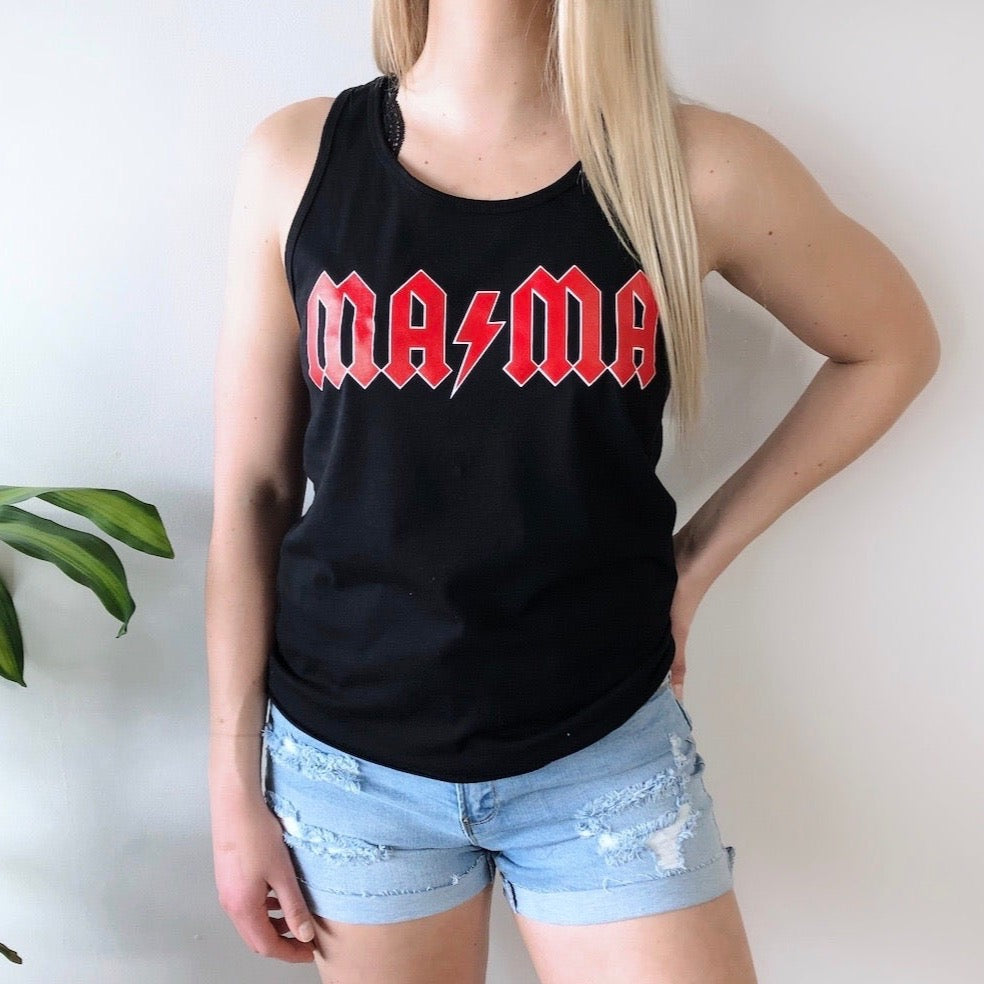 A woman with long blonde hair wears the Rocker MAMA • Unisex Tank Top, featuring bold red AC/DC-inspired MAMA text, paired with light blue ripped shorts. She stands by a green plant. Bella/Canvas unisex fit ensures ultimate comfort.