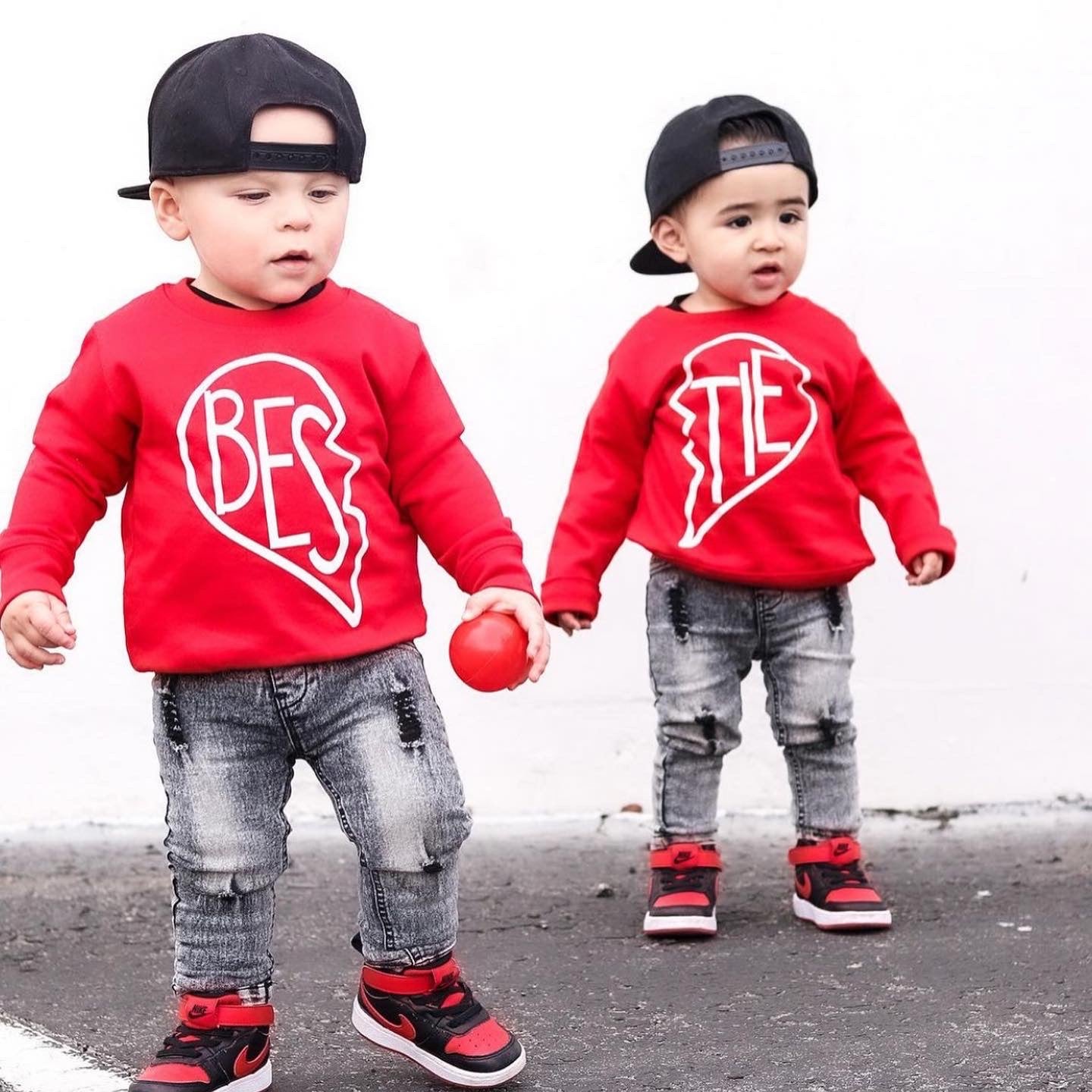 Two toddlers in matching red Bestie Pullover • Red tops, jeans, red-and-black sneakers, and black backwards caps stand outside. Together, their youth pullovers spell “BESTIES.” One holds a red ball.