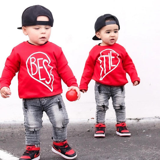 Two toddlers in matching red Bestie Pullover • Red tops, jeans, red-and-black sneakers, and black backwards caps stand outside. Together, their youth pullovers spell “BESTIES.” One holds a red ball.
