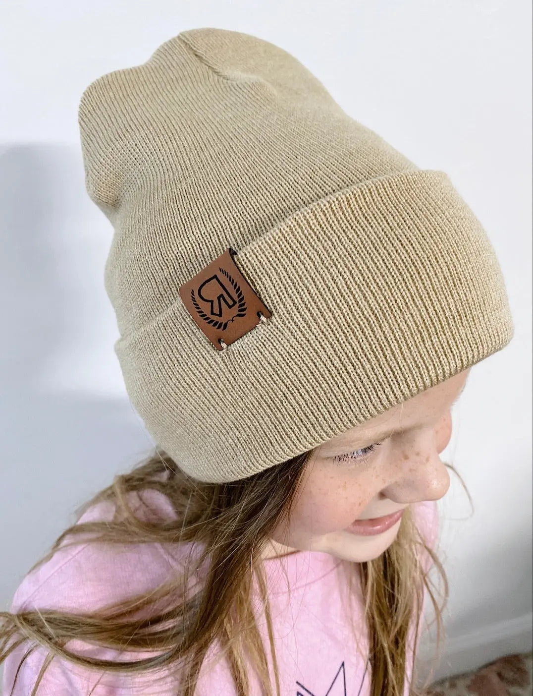 A smiling child with long light brown hair wears a beige Pom Collection Beanie featuring a hand-sewn leather logo patch, paired with a light pink shirt and standing against a white wall.