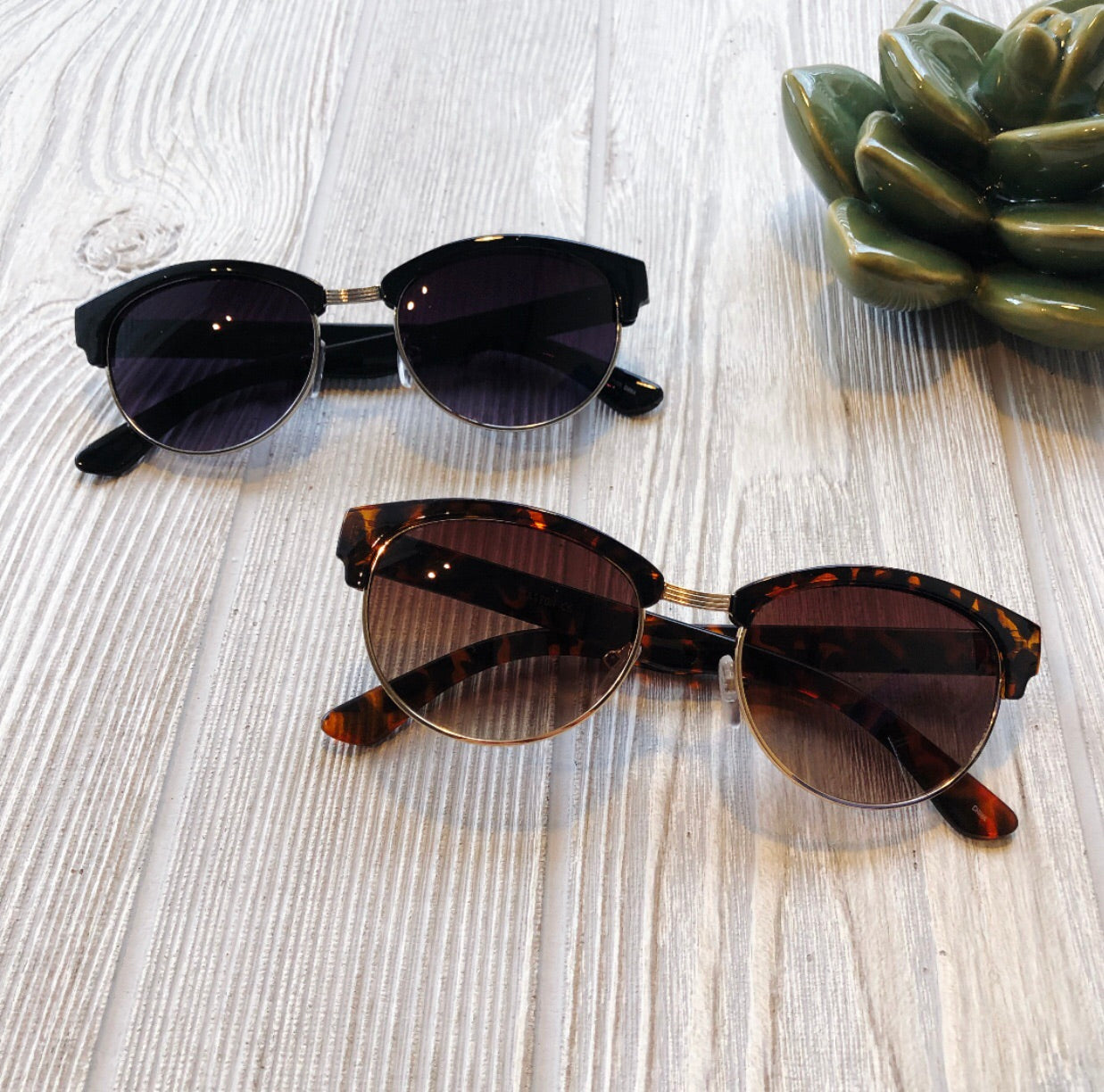 Two pairs of Classic Wayfarer Sunglasses • Adult—one black, one tortoiseshell—feature metal bridges and UV400 protection. Resting on a wooden surface beside a green ceramic succulent, they offer style and safety for every family member.