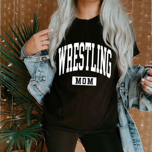 A person with long, wavy light hair wears the Wrestling Mom • Tee with a denim jacket and black pants. Their face is hidden as they stand in front of a plant and string lights, highlighting the unisex fit.