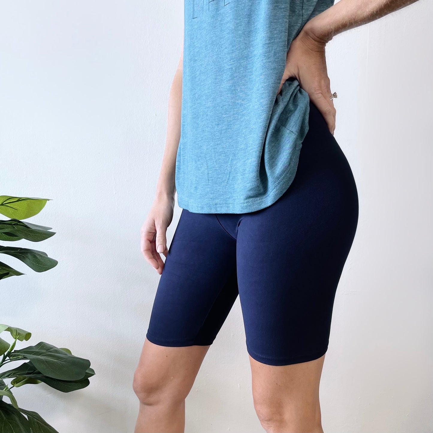 Wearing Navy • Brushed Biker Shorts, a person stands with one hand on their hip against a white wall, while a green potted plant is partially visible in the corner.