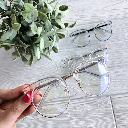 A hand with pink nails holds Kayla Blue Light Glasses in clear rims above a light wooden surface, with two more pairs of Kayla Blue Light Glasses and a green potted plant in the background—perfect for reducing headaches and eye strain.