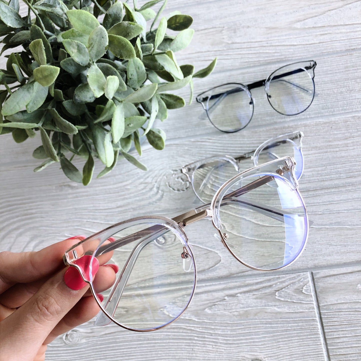 A hand with pink nails holds Kayla Blue Light Glasses in clear rims above a light wooden surface, with two more pairs of Kayla Blue Light Glasses and a green potted plant in the background—perfect for reducing headaches and eye strain.