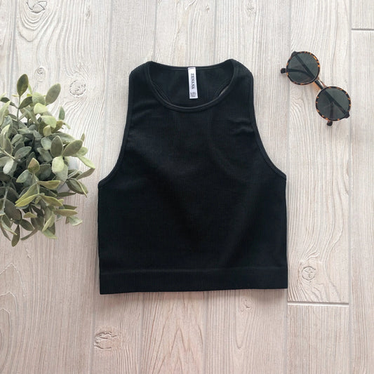 The Sophia High-Neck Crop in black is displayed flat on a light wooden surface, accompanied by a small green potted plant and tortoiseshell sunglasses.