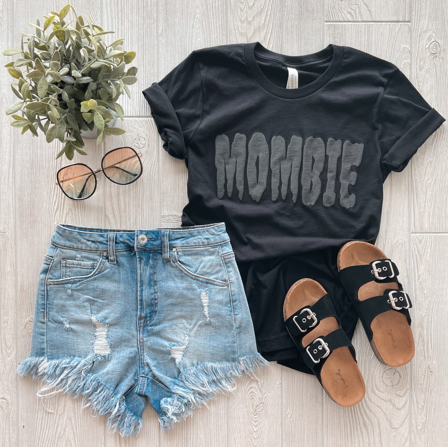 Flat lay of the MOMBIE • Black Tonal Tee with a unisex fit, styled with distressed denim shorts, black buckled sandals, round sunglasses, and a small potted plant on a light wooden floor.