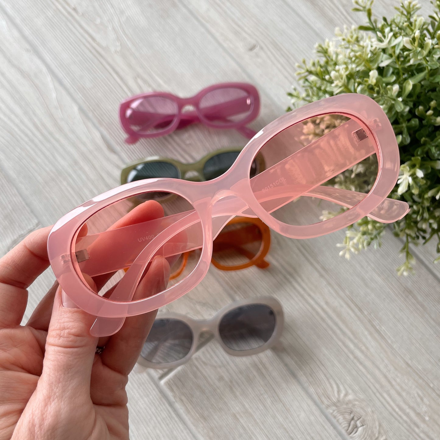 A hand holds oversized pink Poppy Sunglasses (UV protected) above a light wooden surface, with four other Poppy Sunglasses for adults—purple, green, orange, and white—displayed nearby. More colors available! Final Sale Item.