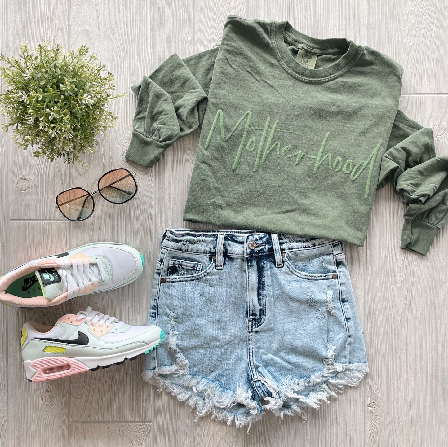 An olive Motherhood • Olive Long Sleeve in soft ring-spun cotton, distressed denim shorts, pastel Nike sneakers, round sunglasses, and a small green plant are arranged on a light wood surface.