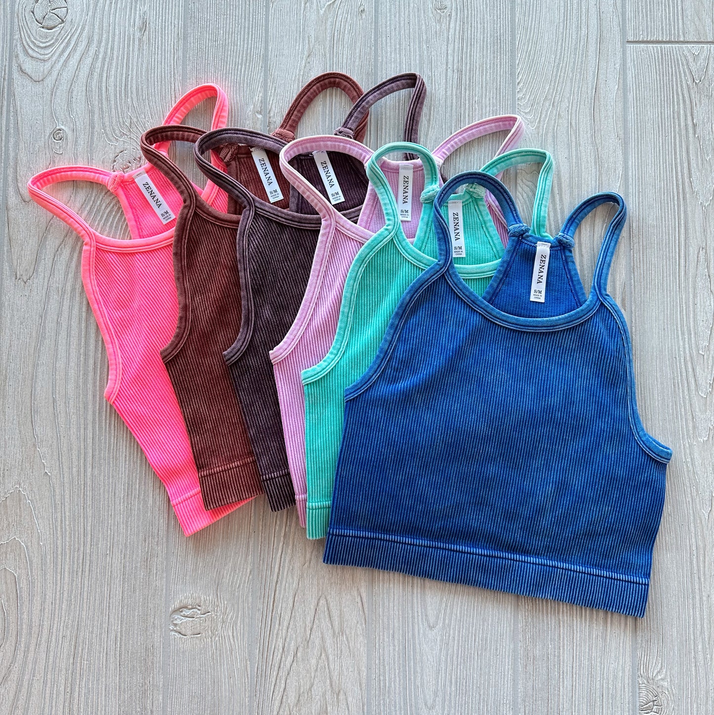 Six Luna Vintage Crop tops from the Tampa Collection—pink, maroon, brown, mint green, teal, and navy blue—are displayed in a fan arrangement on a light wooden floor. These high neck ribbed tanks are stretchy for comfort and easy layering.