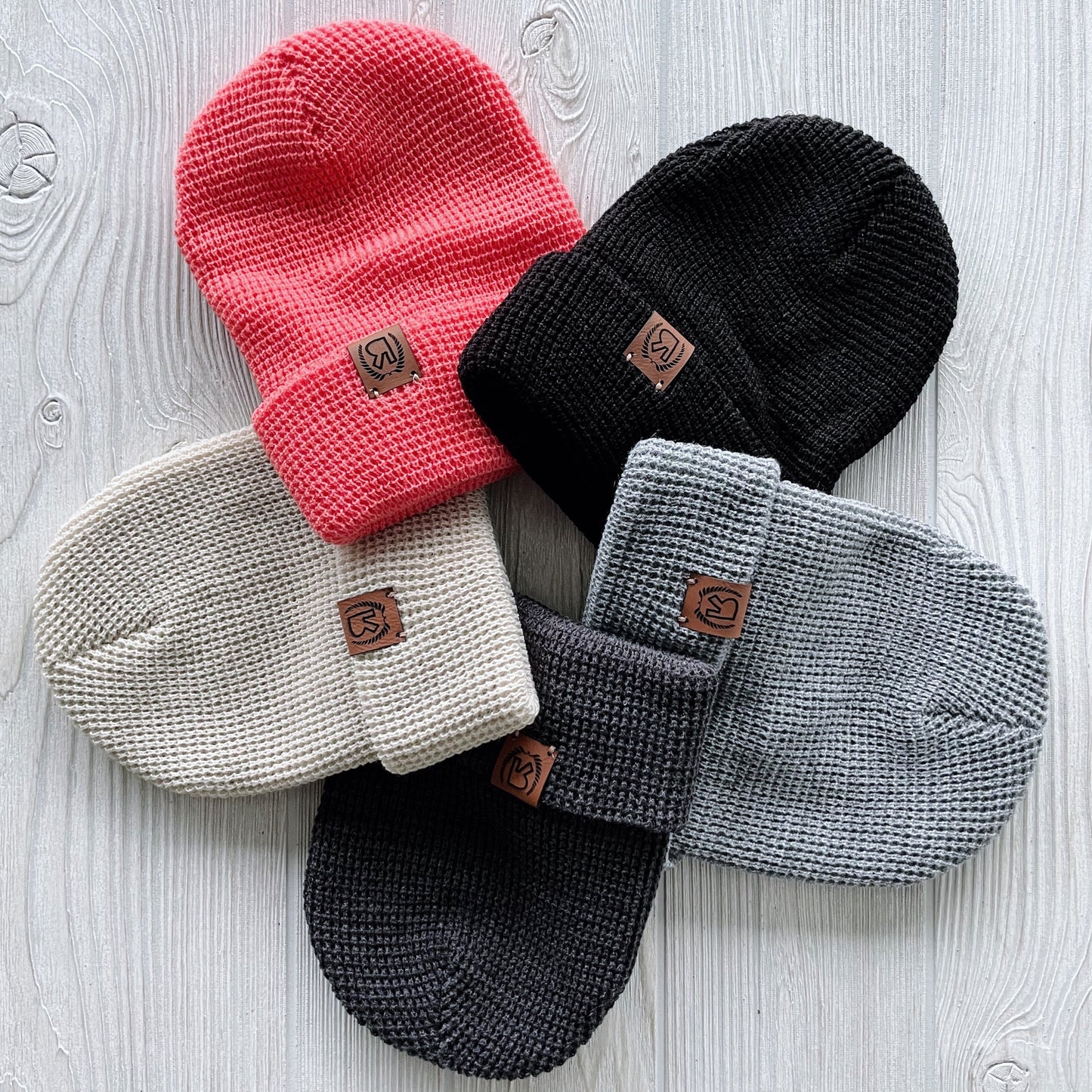 The Waffle Beanie Collection features five winter beanies—in pink, black, off-white, dark gray, and light gray—arranged in a circle on a light wooden surface, each with a brown logo patch and adjustable cuff for the perfect fit.