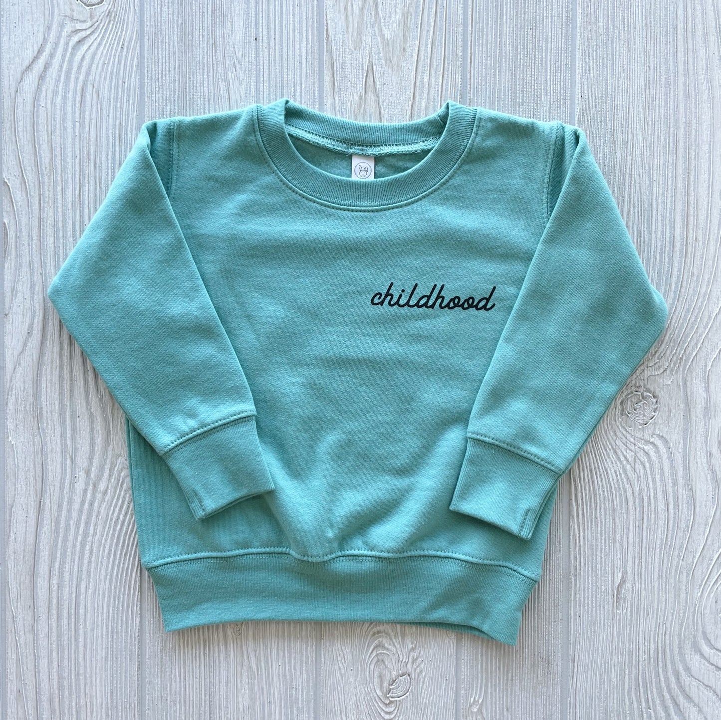 The Childhood • Sage Pullover is a sage green oversized long-sleeve sweatshirt with childhood embroidered in black cursive on the left chest, shown laid flat on a light wood surface.