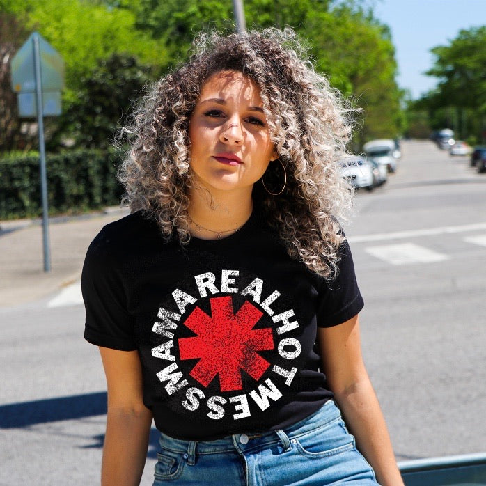 A woman with curly hair stands outdoors in the sun wearing the REAL HOT MESS MAMA black tee, inspired by the Red Hot Chili Peppers, featuring “REAL HOT MAMA MESS” text and a red asterisk. Trees, cars, and a street are visible behind her.