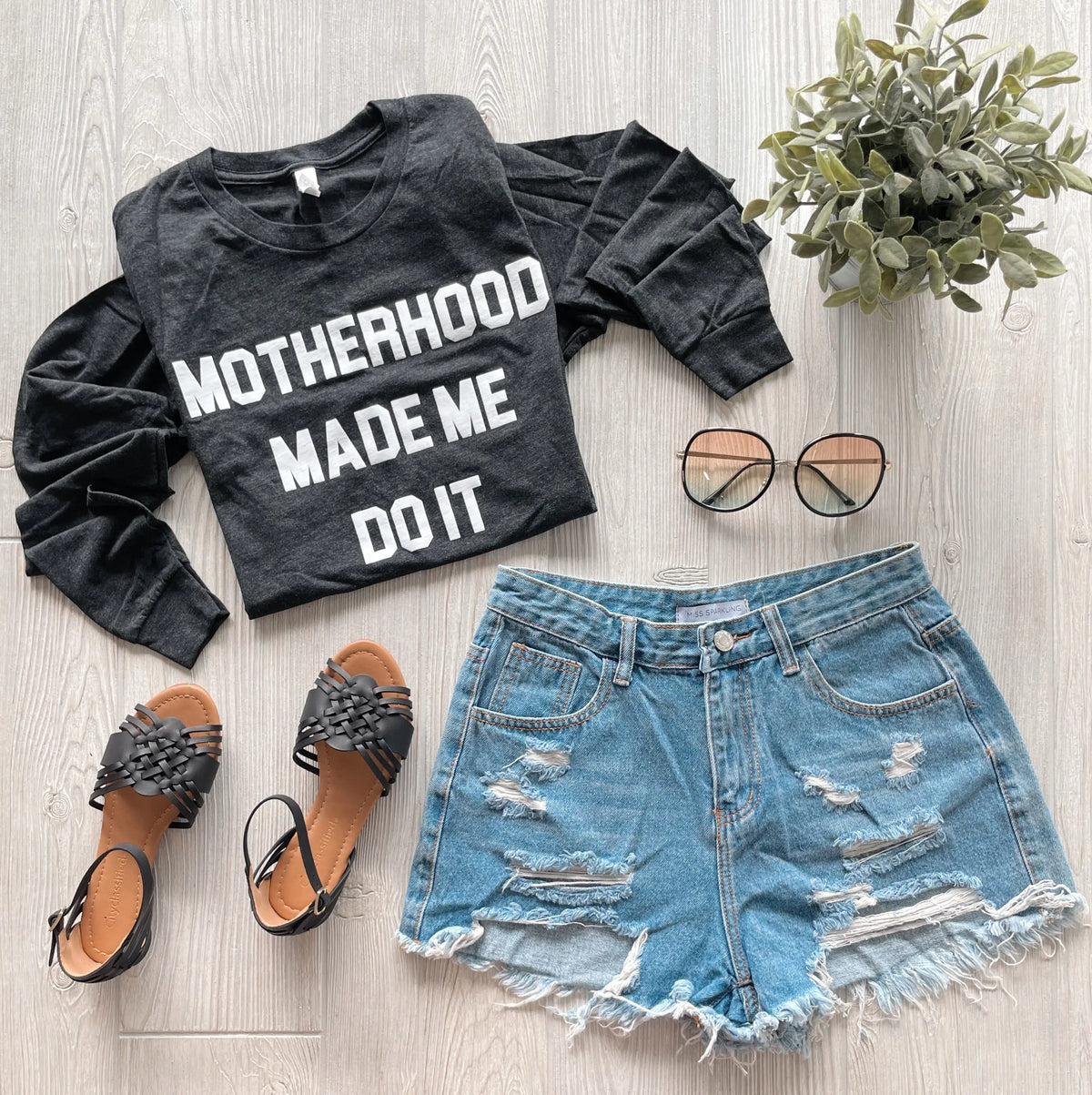 A flat lay features the Motherhood Made Me Heather Black Tri-blend Long Sleeve, distressed denim shorts, black woven sandals, round sunglasses, and a potted plant on a light wood surface.