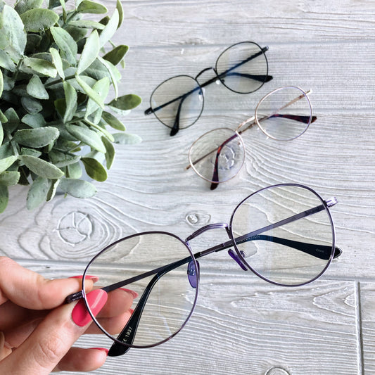 A hand with pink nail polish holds Demi Blue Light Glasses in purple, designed to reduce headaches and eye strain, over a light wooden table. Two more pairs of round glasses and a green potted plant are also on the table. Available in more colors!.