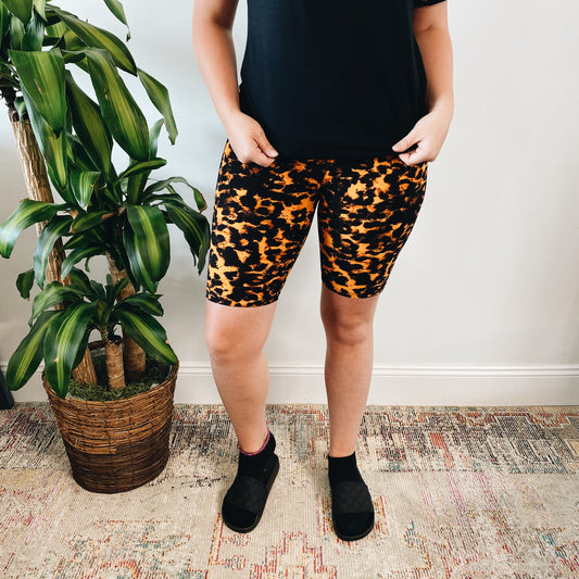 Only the lower half of a person is visible indoors, standing on a patterned rug near a potted plant while wearing Marbled Cheetah • Brushed Biker Shorts.