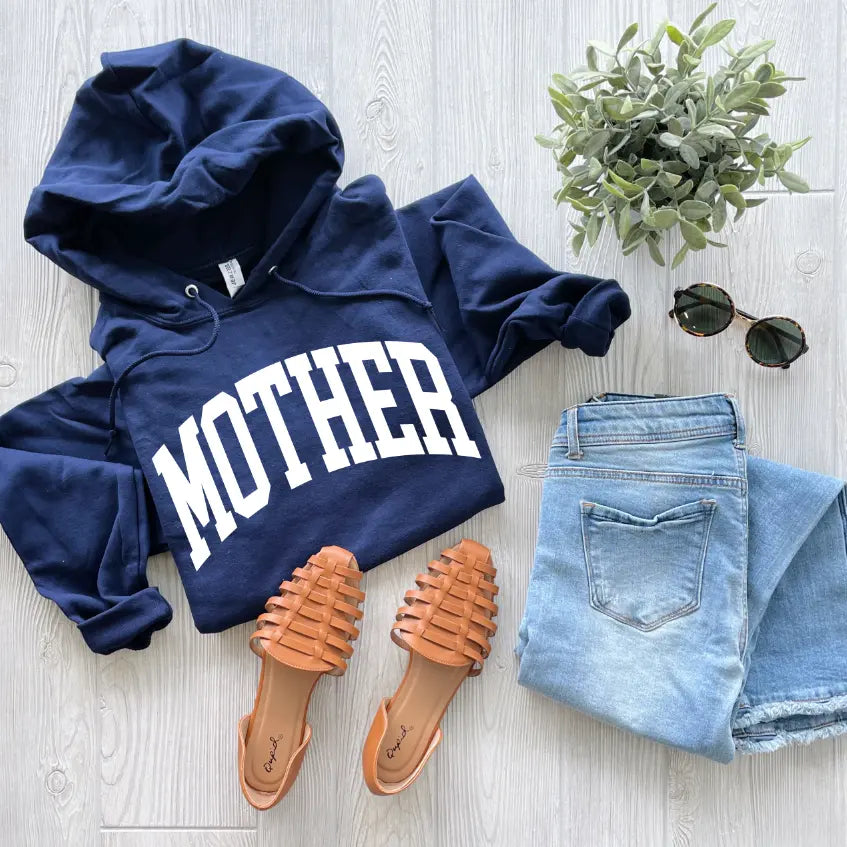 A MOTHER navy hoodie, light blue jeans, tan woven sandals, round sunglasses, and a potted plant are arranged on a light wood floor.