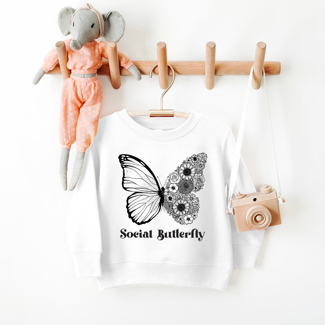 The Social Butterfly • Kids Pullover is a white, oversized fit sweatshirt with a butterfly graphic—one wing floral and Social Butterfly text. Displayed on a wooden rack with a fabric elephant doll and toy camera. Unisex sizing available.