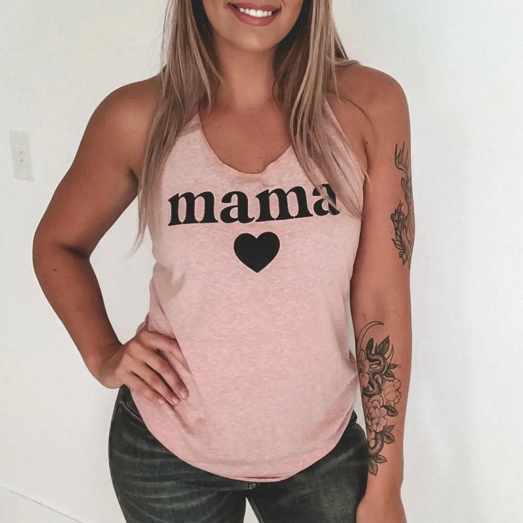 Smiling woman in black jeans shows a floral forearm tattoo while wearing the New Mama Heart • Desert Pink Tank, a women’s racerback top featuring a mama design with heart print.