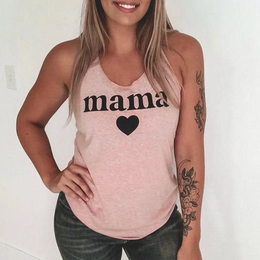 Smiling woman in black jeans shows a floral forearm tattoo while wearing the New Mama Heart • Desert Pink Tank, a women’s racerback top featuring a mama design with heart print.