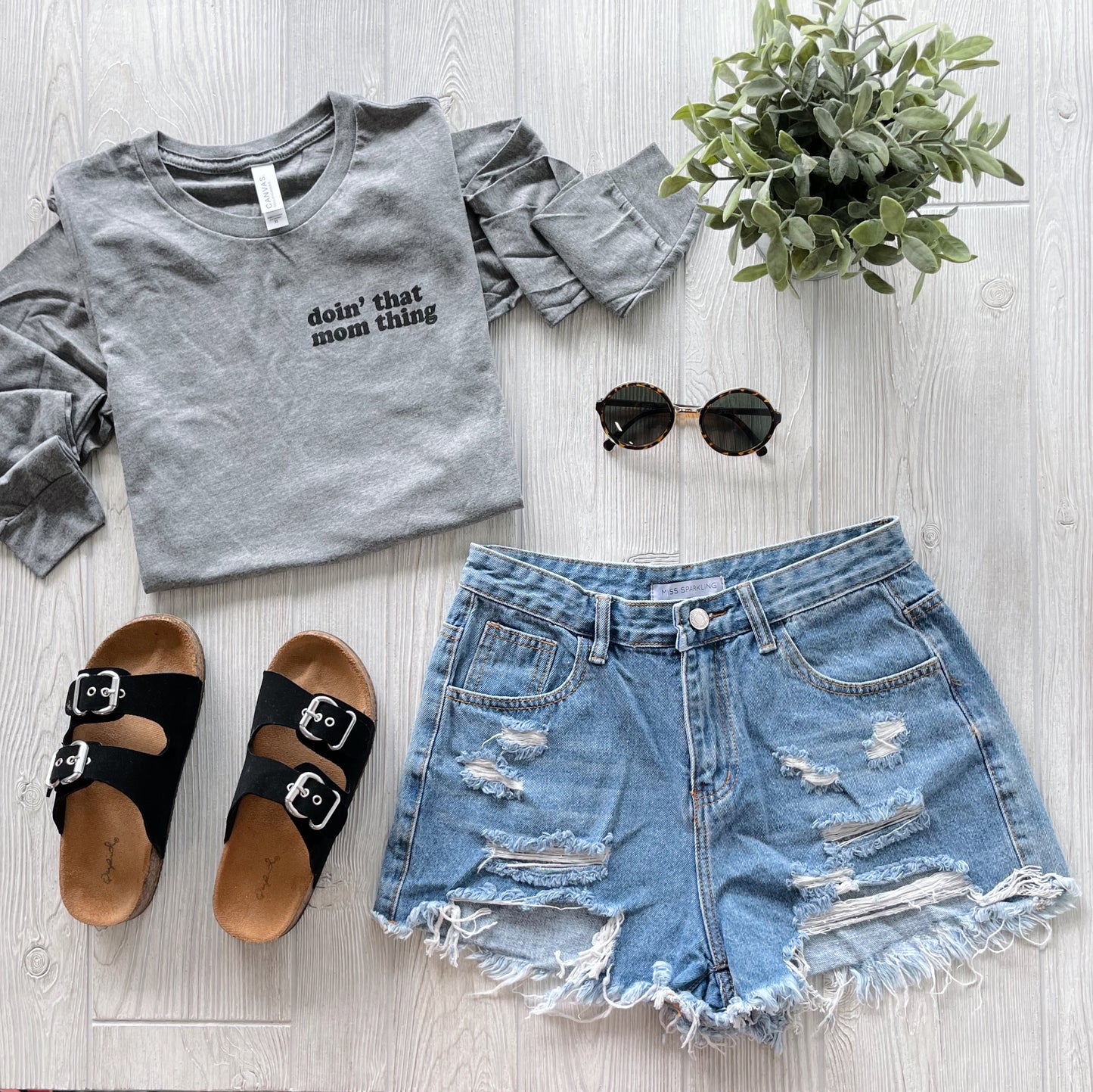 Flat lay of a casual outfit featuring the Mom Thing • Long Sleeved gray triblend tee in unisex sizing with “doin’ that mom thing” text, paired with distressed denim shorts, black sandals, round sunglasses, and a small potted plant on light wood.