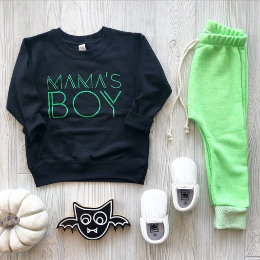 Flat lay of a child’s outfit: Mamas Boy Pullover • Black & Lime, neon green sweatpants, white baby shoes, a white pumpkin, and a black bat decoration on a light wood background.
