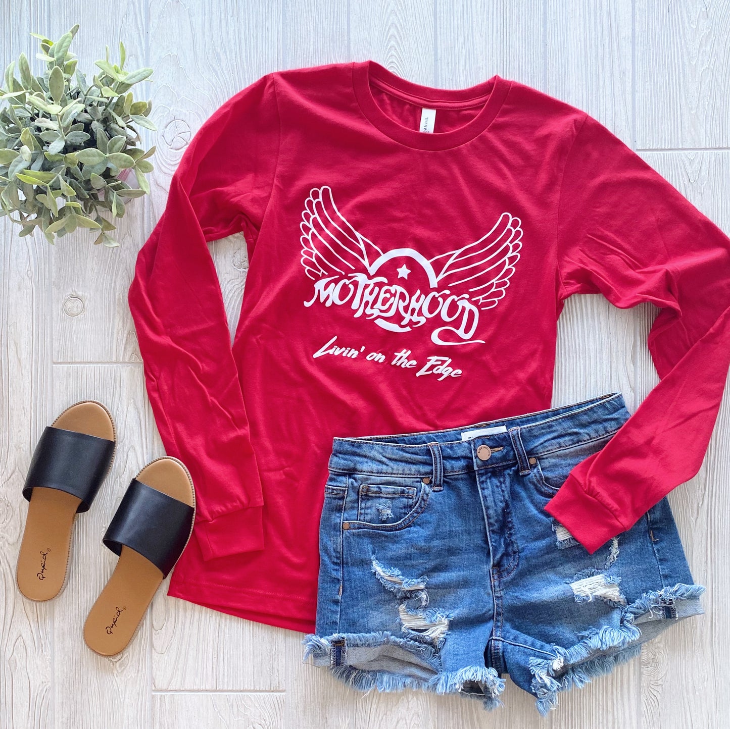 A flat lay of the red Aerosmith Mama • Long Sleeved tee featuring Motherhood Livin on the Edge with a winged heart graphic, paired with distressed denim shorts, black slides, and a potted plant on a light wood floor.