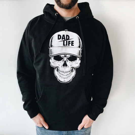 Wearing the Dad Life Skeleton • Black Hoodie, made from soft ring-spun cotton, featuring a bold grinning skull in a DAD LIFE cap. The hoodie has a spacious front pocket for resting hands.