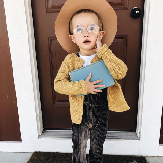 A young child wearing Lennon Sunnies • Kids, a wide-brimmed tan hat, mustard yellow cardigan, and striped pants stands before a brown door, holding a closed blue book and looking up in surprise.