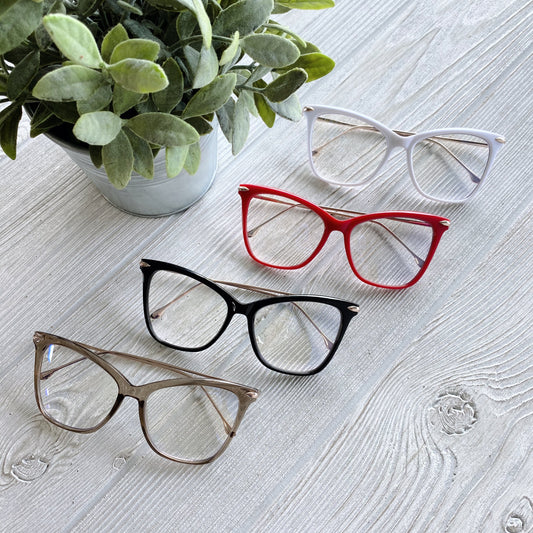 Four Chloe Blue Light Glasses—white, red, black, and beige—are displayed on a light wooden surface beside a potted plant, offering stylish options for eye strain reduction. Available in more colors!.