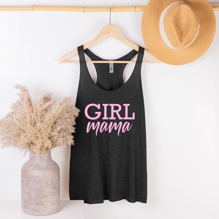 The New Girl Mama • Charcoal Tank, a dark gray racerback with “Girl Mama” in pink, hangs stylishly on a wooden hanger beside a beige vase of pampas grass and a tan straw hat against a white wall.