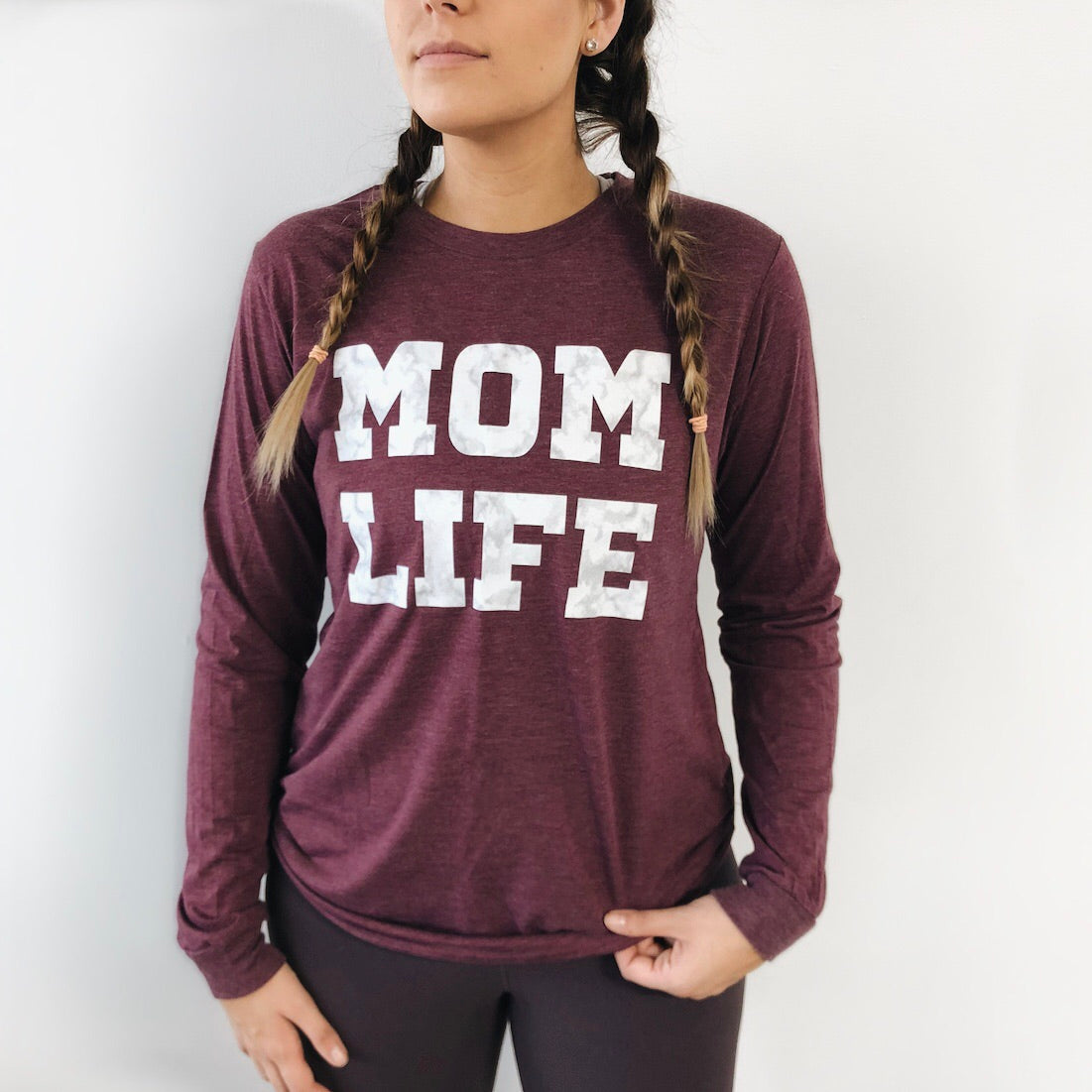 A woman with braided hair wears the MOM LIFE Marble • Heather Maroon Long Sleeve, featuring bold white MOM LIFE letters. Unisex sizing ensures comfort for all, shown here against a plain white background.