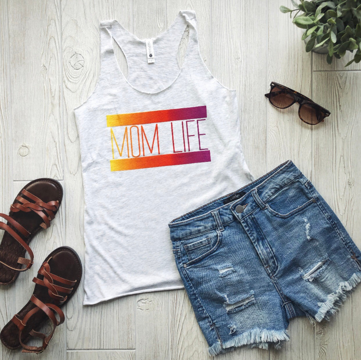 Flat lay of the Mom Life Gradient • Ash Tank featuring bold gradient lettering, styled with distressed denim shorts, brown sandals, and sunglasses on a light wood floor. A potted plant in the corner adds a fresh accent.