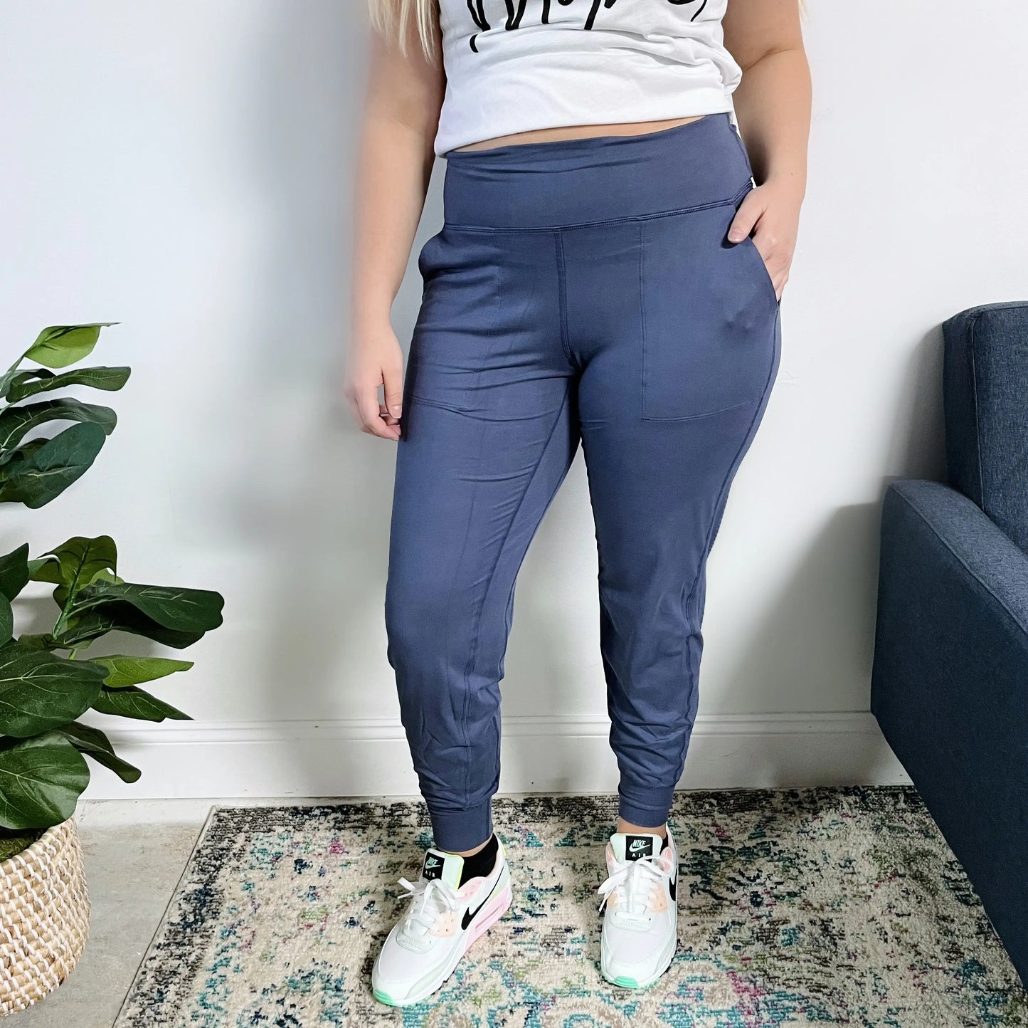 A person wearing Lexi Yoga Pant in Denim Blue, a white graphic t-shirt, and white sneakers stands indoors on a patterned rug near a green potted plant and blue sofa. Their face is not visible.