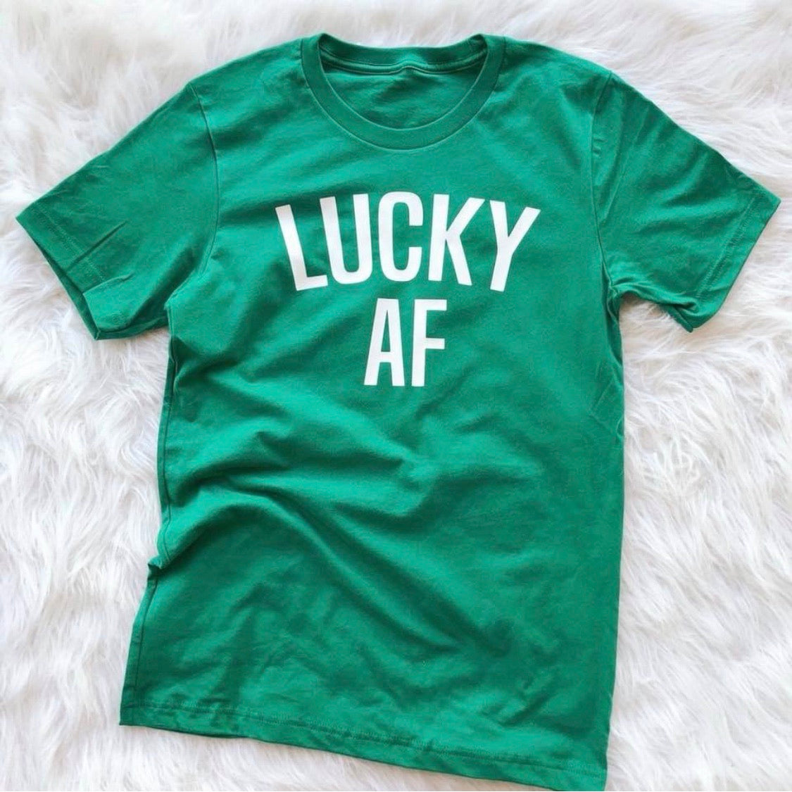 The Lucky (and fun) • Adult Tee is a kelly green shirt with “LUCKY AF” in bold white letters, shown laid flat on a fluffy white background. Unisex sizing ensures a comfortable fit for everyone.