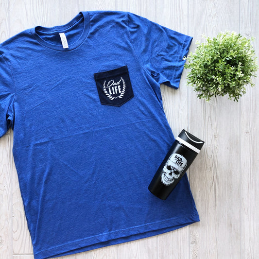 A Blue Dad Life • Pocket Tee made from soft combed, ring-spun cotton with a pocket logo is paired with a black travel mug featuring a skull and Dad Life design, displayed next to a green potted plant on a light wood surface.