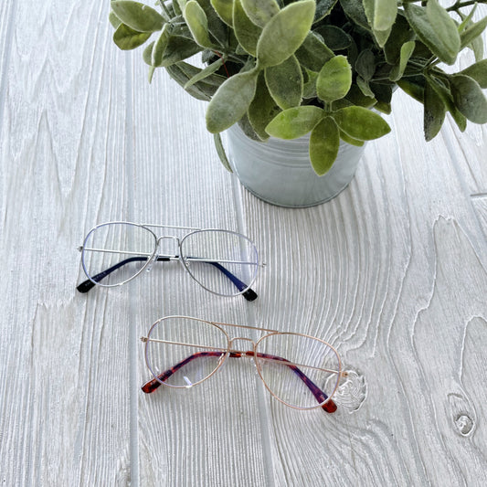 Two pairs of Kids Alex • Blue Light Glasses, one with silver frames and one with rose gold frames, rest on a light wooden surface beside a green leafy plant—perfect for helping kids reduce eye strain.
