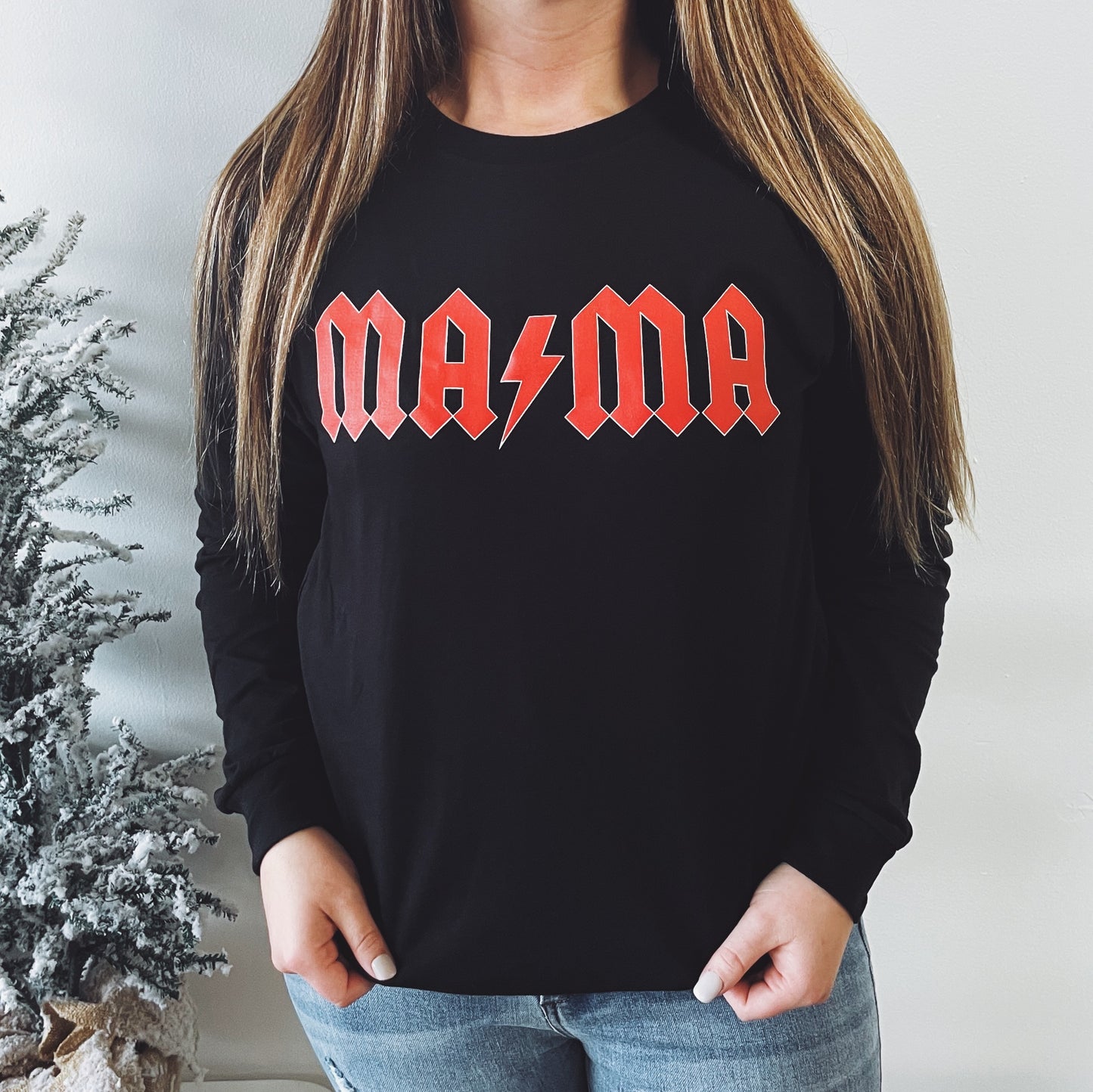 A woman with long straight hair and blue jeans stands against a white wall, wearing the AC/DC Mama • Long Sleeve featuring a bold red MAMA and lightning bolt graphic; a frosted plant is partially visible beside her.