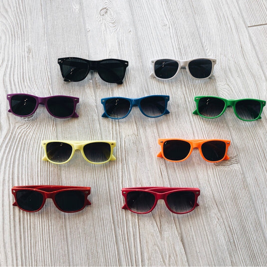 Nine pairs of Classic Sunnies • Kids, each in a different shade—black, white, purple, blue, green, yellow, orange, and pink—are displayed in rows on a light wood surface. Trendy and colorful accessories for any stylish babe.