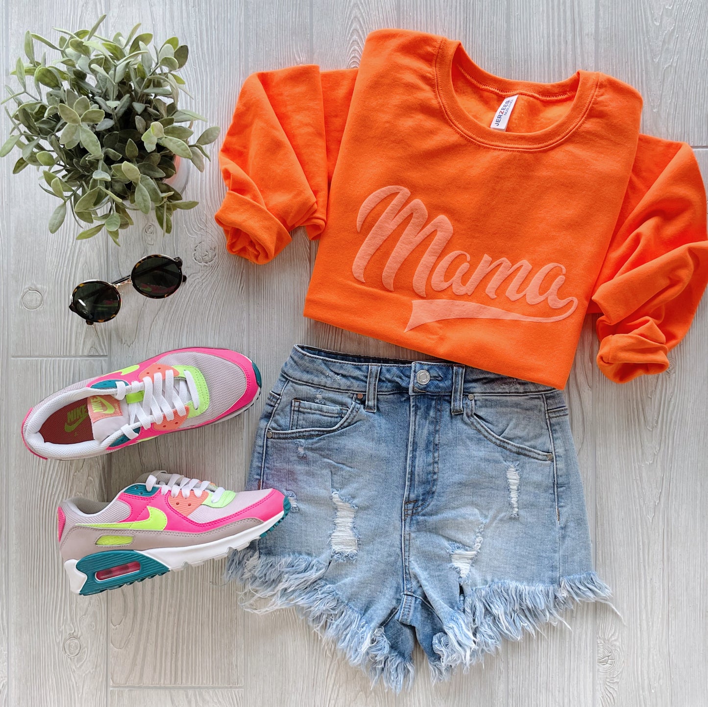 The Sporty Mama • Neon Tonal Pullover, distressed denim shorts, pink and white Nike sneakers, round sunglasses, and a small potted plant are arranged on a light wood background.