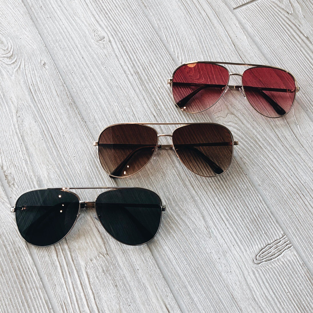 Three pairs of Bri Sunglasses • Adult, each with gold frames and differently tinted lenses (black, brown, red), are arranged diagonally on a light wood surface; all provide reliable UV400 protection.