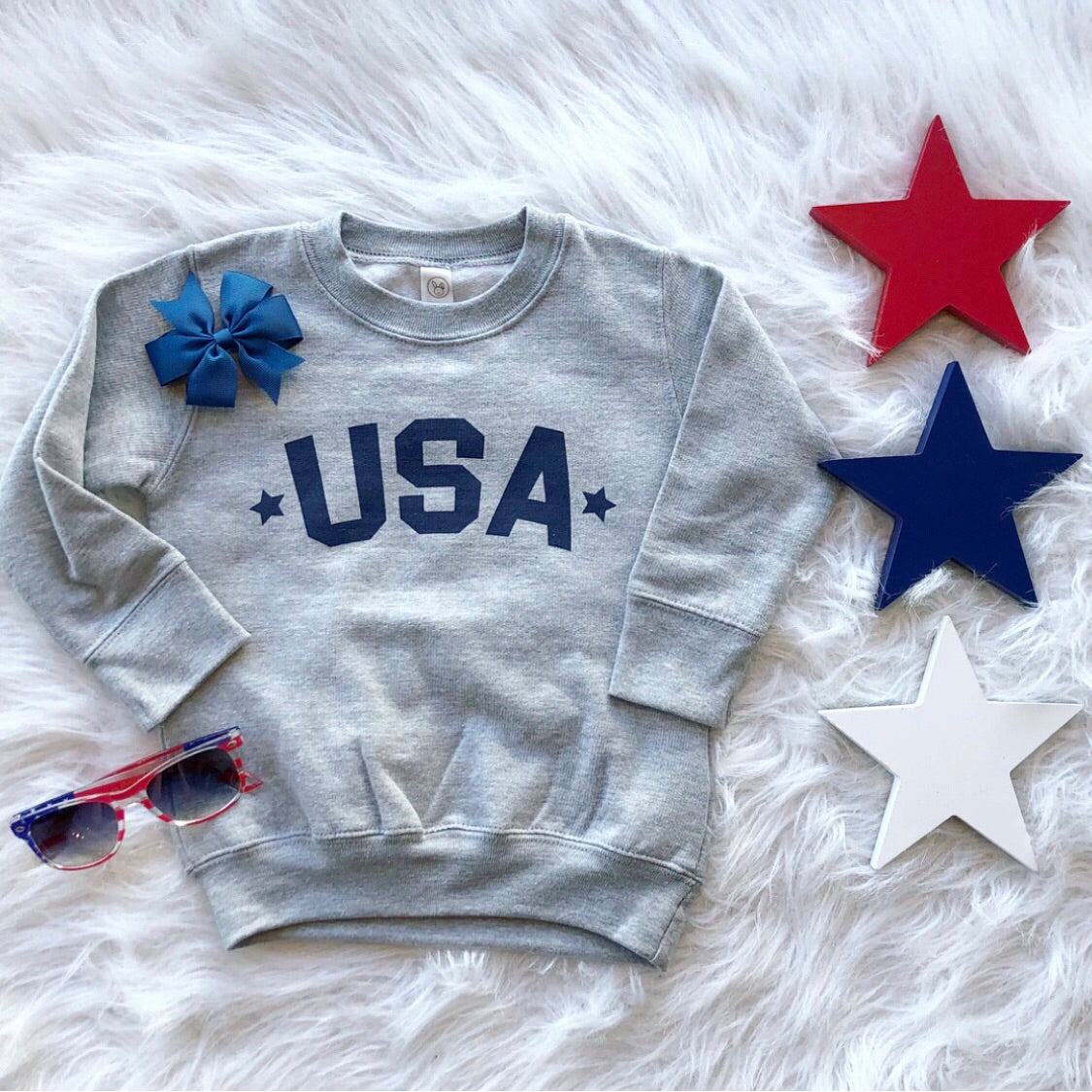 The USA Pullover • Kids! is a gray unisex-fit sweatshirt featuring blue USA text, shown on a white furry surface with red, blue, and white star decorations, a blue bow, and red and blue sunglasses nearby.