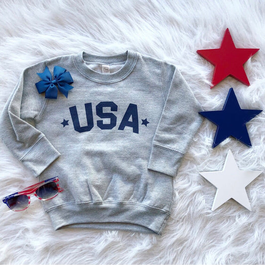 The USA Pullover • Kids! is a gray unisex-fit sweatshirt featuring blue USA text, shown on a white furry surface with red, blue, and white star decorations, a blue bow, and red and blue sunglasses nearby.