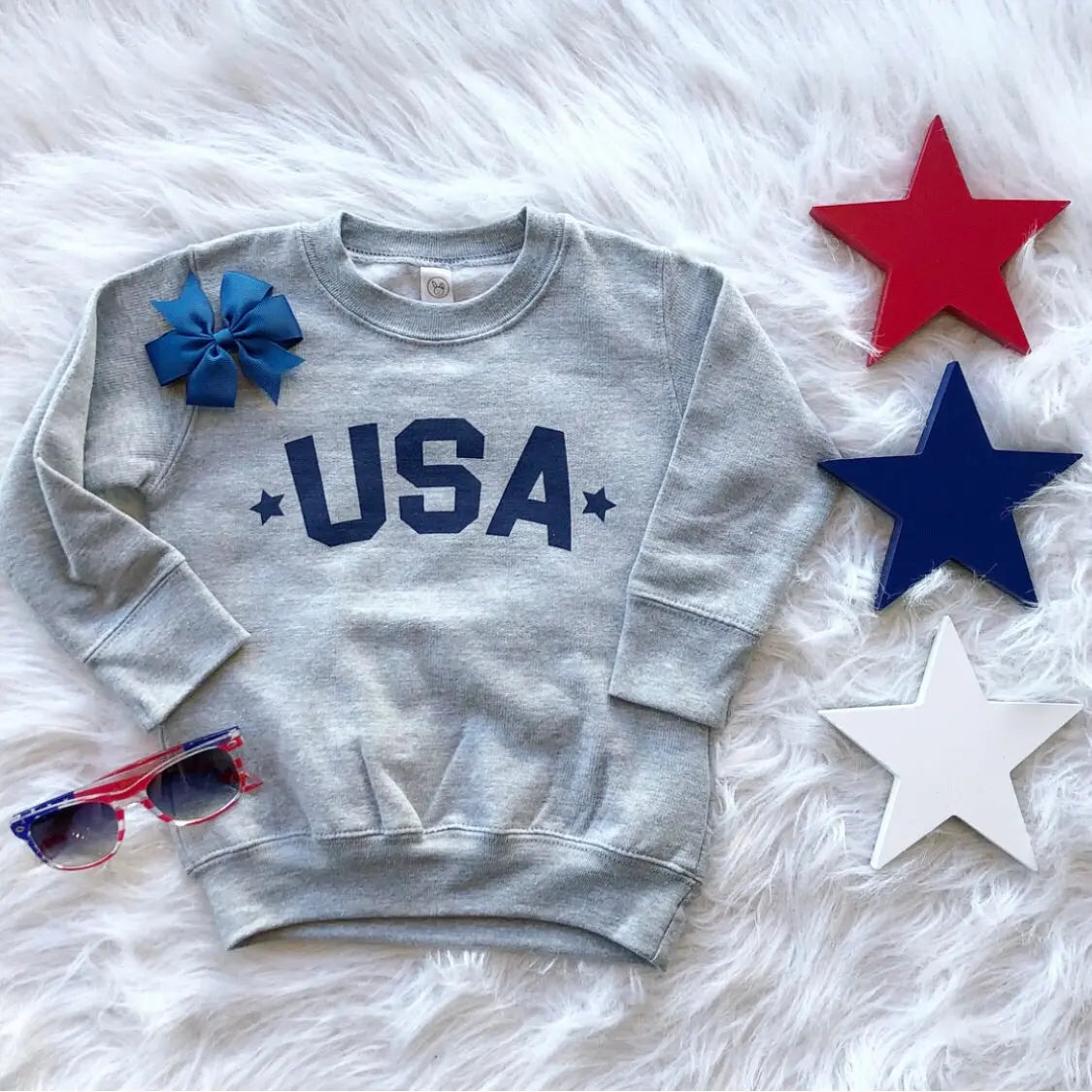 The USA Pullover • Kids! is a gray unisex-fit sweatshirt featuring blue USA text, shown on a white furry surface with red, blue, and white star decorations, a blue bow, and red and blue sunglasses nearby.