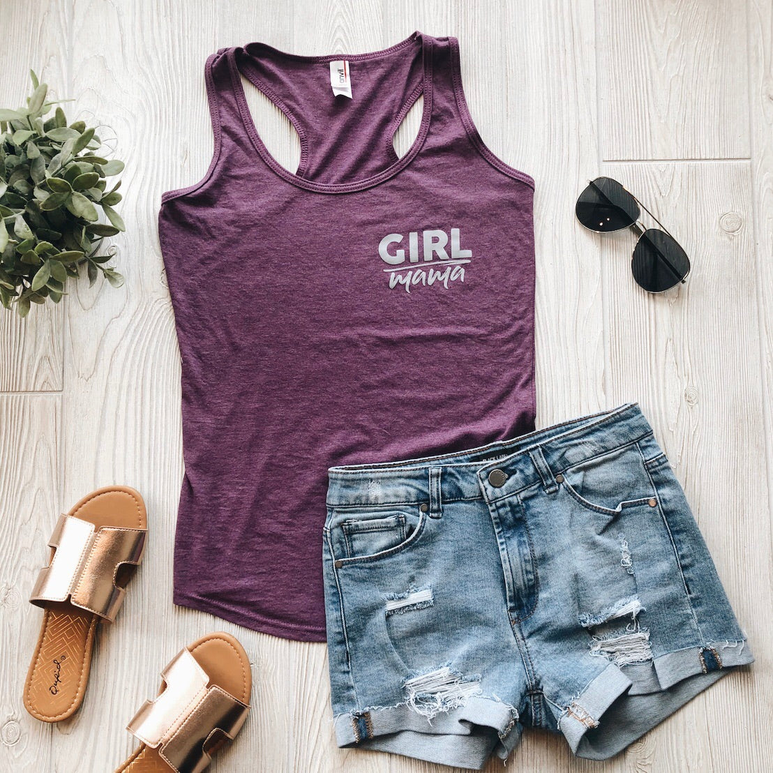 Flat lay of the Girl Mama • Racerback Tank in purple neps jersey, styled with distressed denim shorts, metallic slide sandals, black aviator sunglasses, and a small green plant on a light wooden floor.