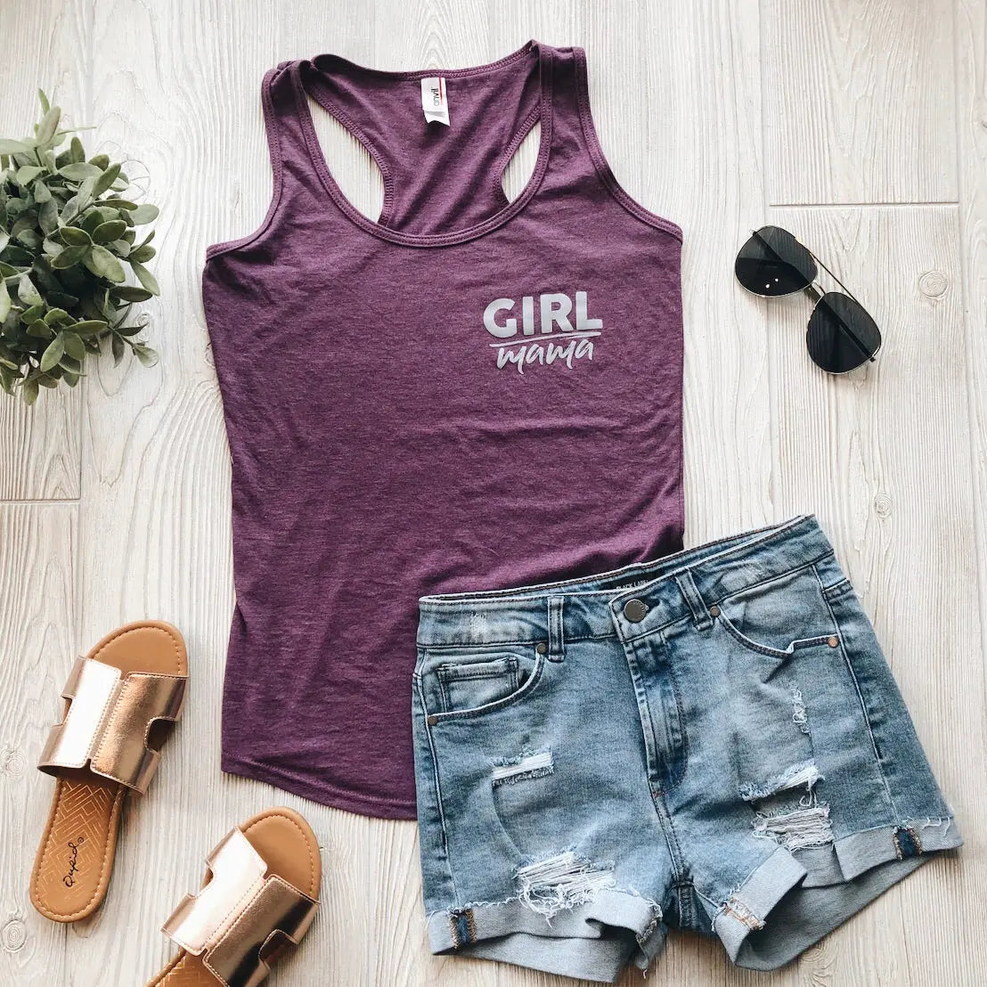 Flat lay of the Girl Mama • Racerback Tank in purple neps jersey, styled with distressed denim shorts, metallic slide sandals, black aviator sunglasses, and a small green plant on a light wooden floor.