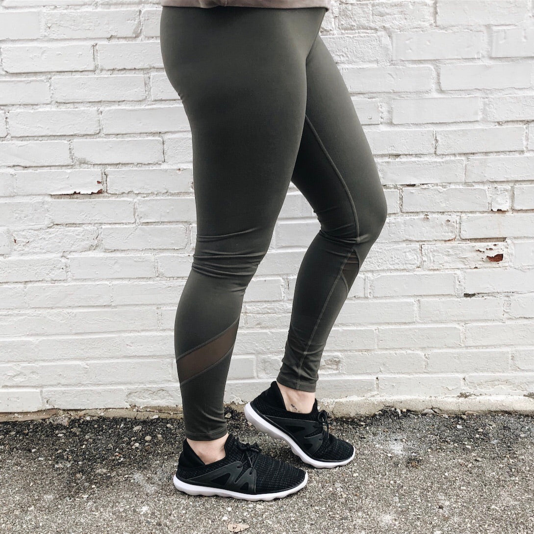 A person models the Mesh Splice Leggings • 1324, featuring an olive green hue and athletic details, with black sneakers. Standing on rough pavement before a white brick wall, only their legs and the yoga waistband are visible.