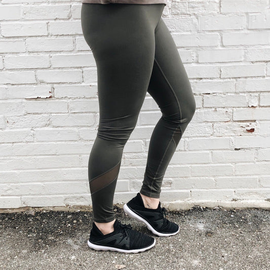 A person models the Mesh Splice Leggings • 1324, featuring an olive green hue and athletic details, with black sneakers. Standing on rough pavement before a white brick wall, only their legs and the yoga waistband are visible.