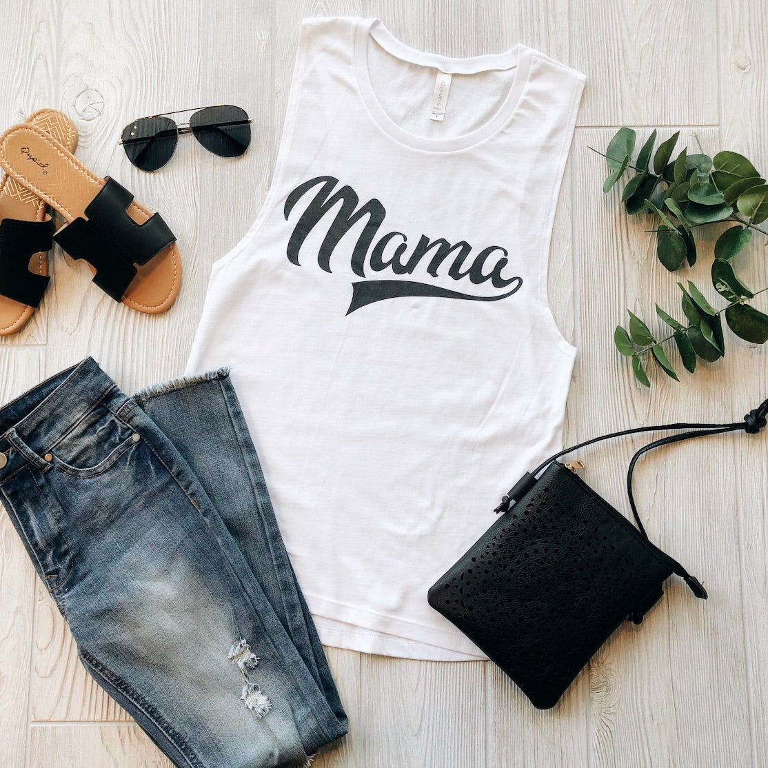 A flat lay featuring the Mama Sporty • White Muscle Tank in white with Mama in black script, styled with distressed blue jeans, black sandals, sunglasses, a crossbody bag, and green leafy sprigs on a wooden surface.